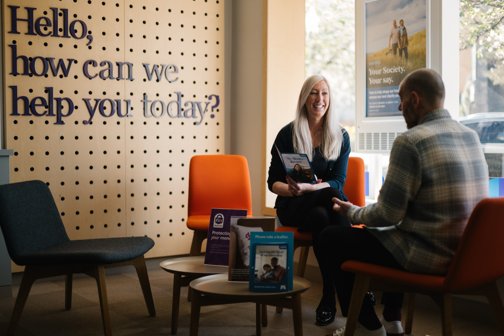 Colleague showing brochure to customer in branch seating area with 'Hello, how can we help you today sign' in the background.